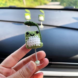 May include: A handmade crochet frog car accessory. The frog is green and white with black eyes and a leaf. It is attached to a spring and suction cup for easy placement on a car dashboard. The text "XINHNV" is visible.