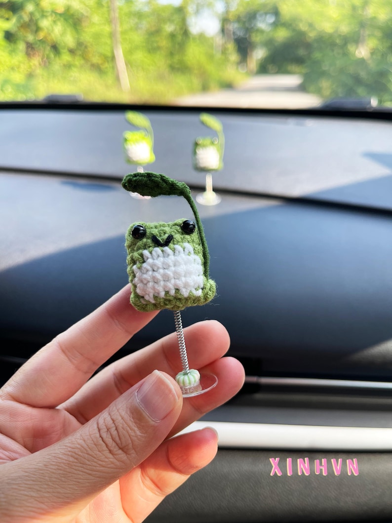 May include: A handmade crochet frog car accessory. The frog is green and white with black eyes and a leaf. It is attached to a spring and suction cup for easy placement on a car dashboard. The text "XINHNV" is visible.