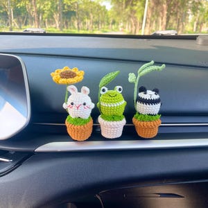 May include: Three crocheted plant-themed car accessories. A white bunny with a sunflower, a green frog, and a panda bear, each in a small pot. The accessories are placed on a car dashboard, with a blurred background of trees.