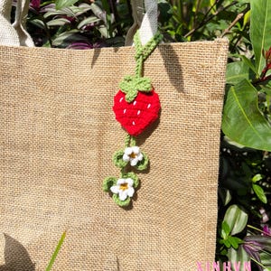 May include: A natural burlap tote bag with cream-colored handles. A crocheted strawberry charm with green leaves and white flowers hangs from the handle. The strawberry is red with white seed details.