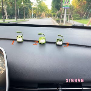 May include: Three handmade crochet frog figurines with green bodies and white bellies, each with a small green leaf. The figurines are numbered 1, 2, and 3. The word "XINHVN" is visible in pink text.