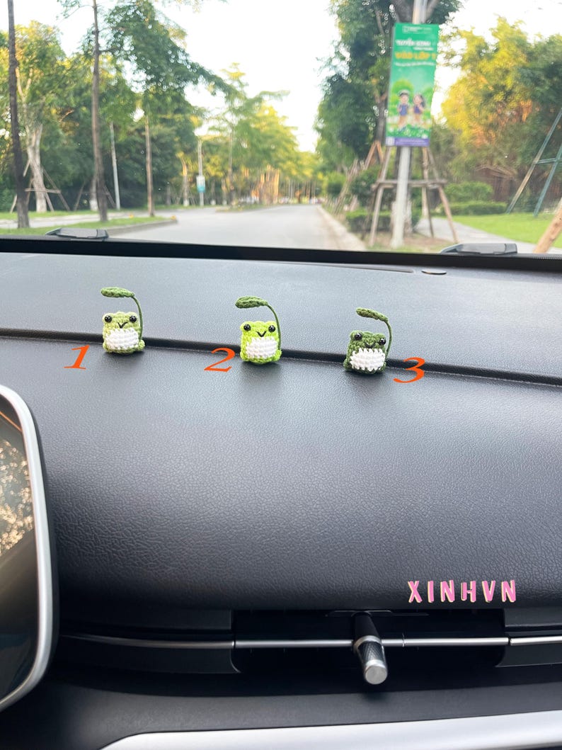 May include: Three handmade crochet frog figurines with green bodies and white bellies, each with a small green leaf. The figurines are numbered 1, 2, and 3. The word "XINHVN" is visible in pink text.