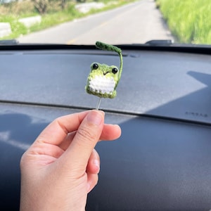May include: A handmade, crocheted frog ornament with a green body, white belly, and black eyes. The frog has a small green leaf attached to its head and is held by a silver wire. The background shows a road and greenery.
