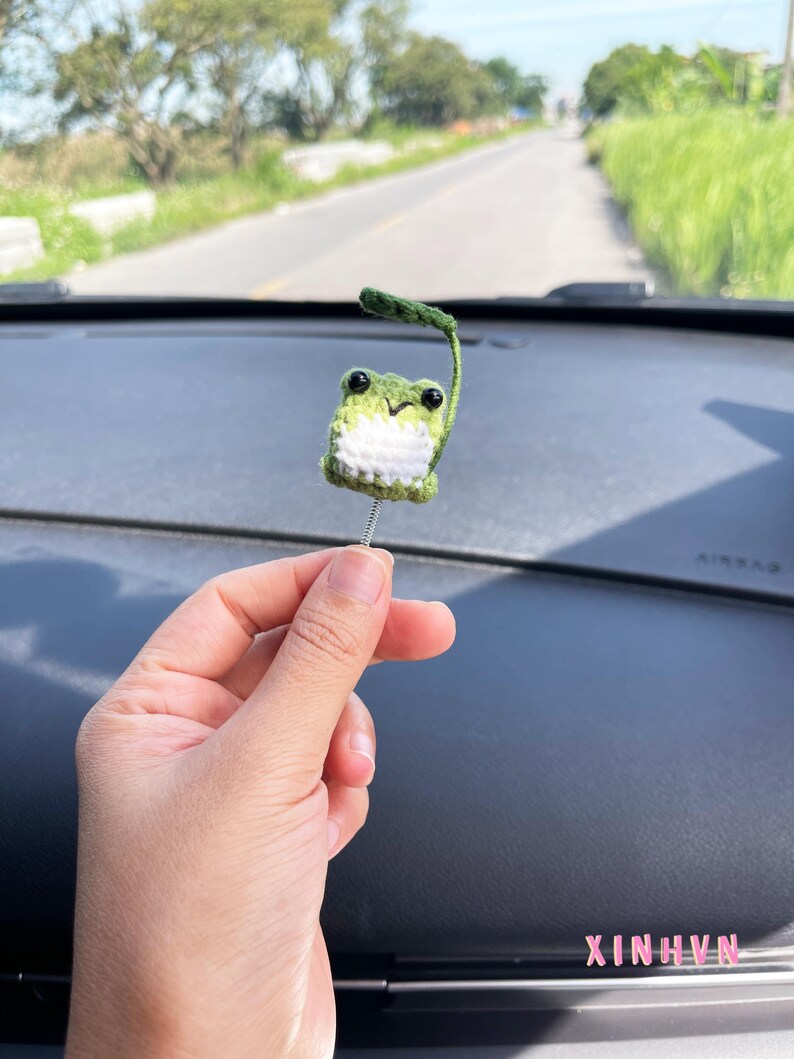 May include: A handmade, crocheted frog ornament with a green body, white belly, and black eyes. The frog has a small green leaf attached to its head and is held by a silver wire. The background shows a road and greenery.