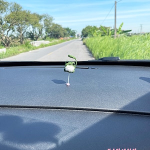 May include: A small, handmade crochet frog car charm. The frog is green and white, with a leaf on its head. The charm is hanging from a pink string. The text "XINHNV" is visible in pink.