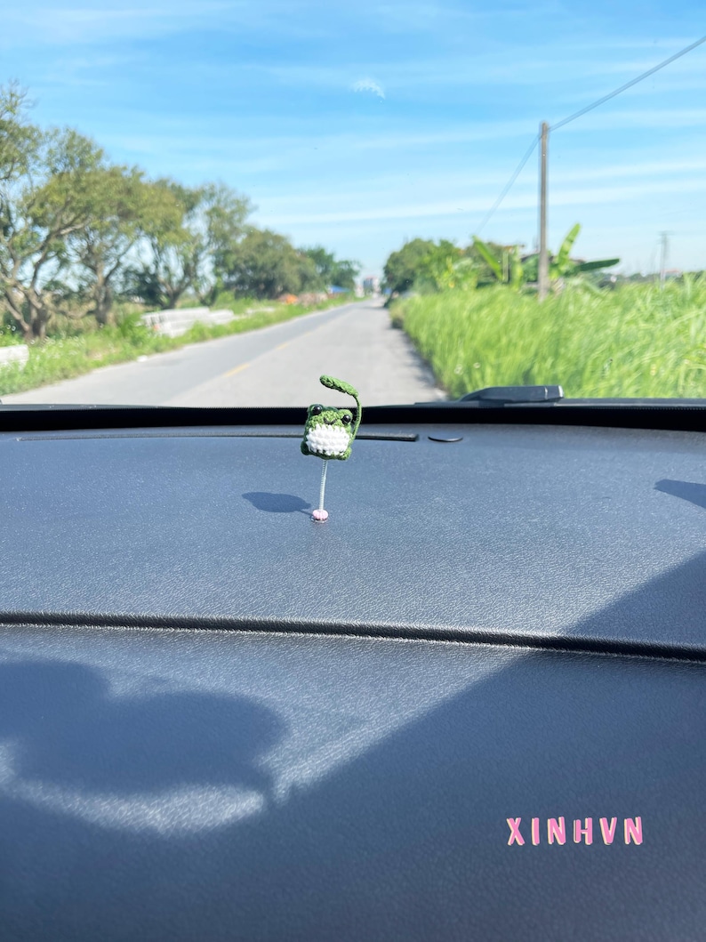 May include: A small, handmade crochet frog car charm. The frog is green and white, with a leaf on its head. The charm is hanging from a pink string. The text "XINHNV" is visible in pink.