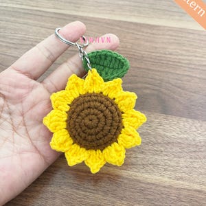 Op de afbeelding: Een gehaakte zonnebloem sleutelhanger met een groen blad. De zonnebloem is geel met een bruin centrum. De sleutelhanger is bevestigd aan een zilveren ring.