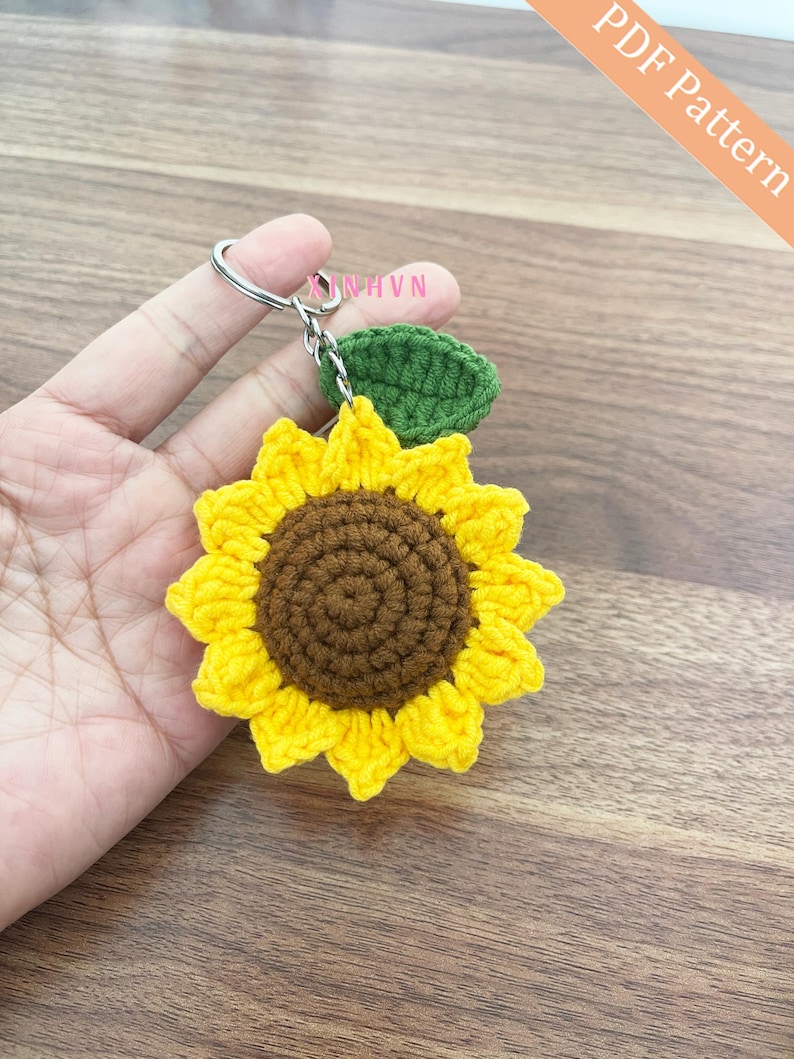 Op de afbeelding: Een gehaakte zonnebloem sleutelhanger met een groen blad. De zonnebloem is geel met een bruin centrum. De sleutelhanger is bevestigd aan een zilveren ring.