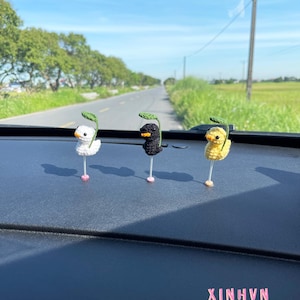 Könnte beinhalten: Drei gehäkelte Entenfiguren in Weiß, Schwarz und Gelb, jede mit einem grünen Blatt. Die Enten befinden sich auf einer dunklen Oberfläche mit dem Wort "XINHVN" in Rosa. Der Hintergrund zeigt eine Straße und einen blauen Himmel.
