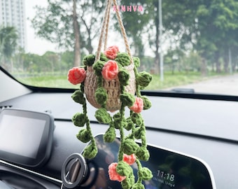 Op de afbeelding: Een gehaakte hangplant met roze bloemen en groene bladeren, hangend aan de achteruitkijkspiegel van een auto. Het dashboard van de auto is zichtbaar op de achtergrond.