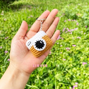 Könnte beinhalten: Ein handgefertigter Schlüsselanhänger in Form einer Kamera. Der Anhänger hat eine weiße Oberseite, einen braunen Körper und ein schwarz-weißes Objektiv. Das Wort "XINGHN" ist sichtbar. Er ist an einem silbernen Schlüsselring befestigt.