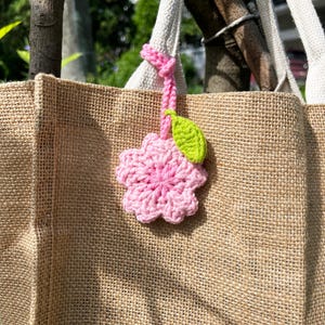 May include: A beige jute tote bag with white handles. A pink crocheted flower with a green leaf charm hangs from the handle. The flower is a light pink colour and the leaf is green. The bag has a woven texture.