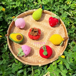 Puede incluir: Una colección de llaveros de frutas hechos a mano con ganchillo, que incluyen una pera, una fresa, un limón, un aguacate y un tomate, dispuestos en una bandeja tejida. Los llaveros son de varios colores, como verde, rojo, amarillo y rosa, sobre un fondo de follaje verde.