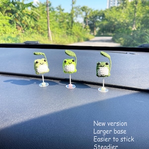 May include: Three handmade crochet frog decorations with green leaves, white bellies, and black eyes. The frogs are mounted on clear stands. Text on the right reads "New version, Larger base, Easier to stick, Steadier."