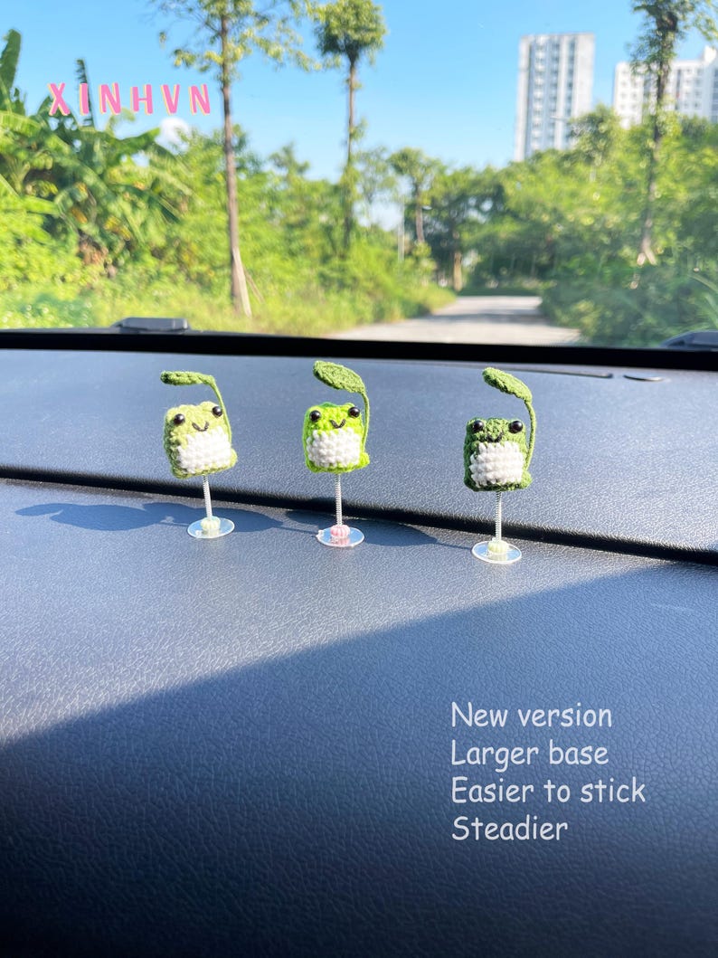 May include: Three handmade crochet frog decorations with green leaves, white bellies, and black eyes. The frogs are mounted on clear stands. Text on the right reads "New version, Larger base, Easier to stick, Steadier."