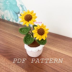 Puede incluir: Una planta de girasol de crochet con dos flores amarillas y hojas verdes en una maceta blanca con una base marrón. La maceta tiene un borde festoneado. La planta está sentada sobre una superficie de madera. El texto "PDF PATTERN" está escrito debajo de la planta.
