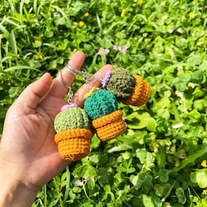 May include: Three crochet cactus keychains in a hand. Each cactus is green or olive green, with a small purple or pink flower, and sits in an orange pot. The keychains are attached to silver metal rings. The background is a field of green grass.