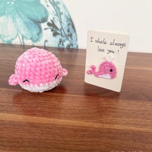 May include: A pink crocheted whale plushie with a white belly and a small white crocheted crown on its head. A card with the text "I whale always love you!" and a small pink crocheted whale on it.