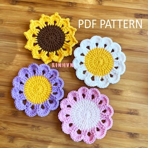 Könnte beinhalten: Vier handgefertigte gehäkelte Blumenuntersetzer in Gelb, Weiß, Lila und Rosa, jeweils mit gelber Mitte. Die Untersetzer sind auf einer Holzoberfläche angeordnet. Der Text "PDF PATTERN" ist sichtbar.