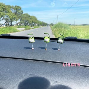 May include: Three handmade crochet frog figurines with green and white bodies, each with a leaf above its head. The frogs are standing on a dark surface, with a road and trees in the background. The word "XINHNIX" is visible.