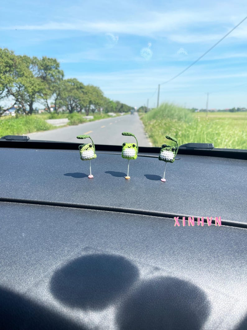 May include: Three handmade crochet frog figurines with green and white bodies, each with a leaf above its head. The frogs are standing on a dark surface, with a road and trees in the background. The word "XINHNIX" is visible.