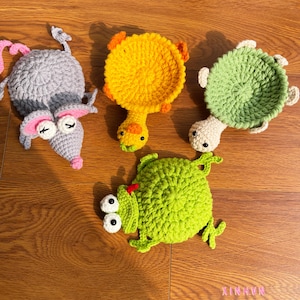 Puede incluir: Juego de posavasos de animales tejidos a ganchillo en gris, amarillo y verde. El conjunto incluye un ratón, un pato, una tortuga y una rana. Los posavasos están hechos a mano y presentan detalles faciales y superficies texturizadas. La palabra "XINHIN" es visible en la esquina inferior derecha.