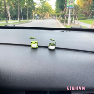 May include: Two small, crocheted frog figurines with green bodies, white bellies, and leaf-like details on their heads. The frogs are placed on a dark dashboard, with the text "XINHVN" in pink. The background shows a road and trees.