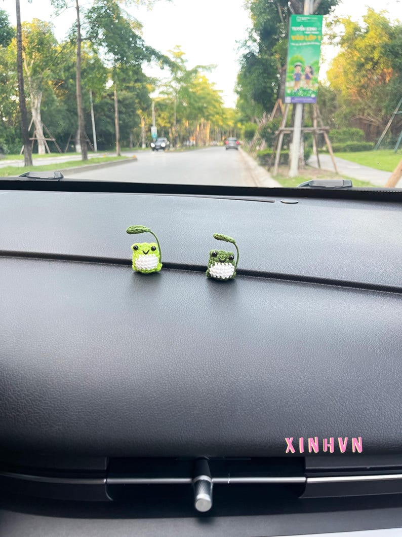 May include: Two small, crocheted frog figurines with green bodies, white bellies, and leaf-like details on their heads. The frogs are placed on a dark dashboard, with the text "XINHVN" in pink. The background shows a road and trees.