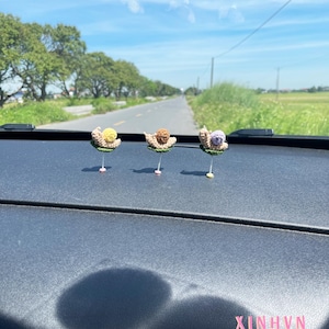 May include: Three handmade crochet snail figurines with yellow, brown, and purple shells are displayed on a dark surface. Each snail is attached to a pin. The background shows a road, trees, and a blue sky.