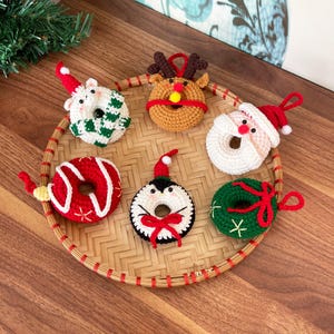 以下が含まれることがあります： クリスマスのキャラクターが描かれたドーナツ型のクロシェ編みオーナメントが6つ。オーナメントは赤、緑、白、茶色、黒です。オーナメントは、竹製の編まれたトレイに並べてあります。