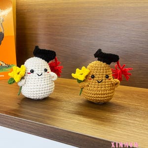 Puede incluir: Dos figuras de ganchillo, una blanca y otra amarilla, cada una con un birrete negro. Sostienen flores amarillas y tienen detalles rojos. Las figuras sonrientes se exhiben en una estantería de madera.