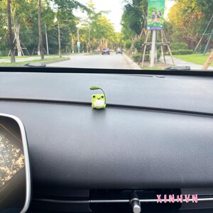 May include: A small, crocheted frog figurine with a leaf on its head sits on a car dashboard. The frog is green and white with a smiling face. The dashboard is black, and the background shows a road with trees and a sign.