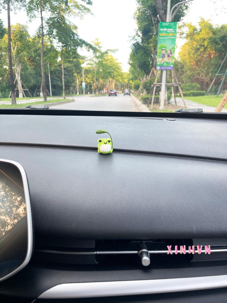May include: A small, crocheted frog figurine with a leaf on its head sits on a car dashboard. The frog is green and white with a smiling face. The dashboard is black, and the background shows a road with trees and a sign.