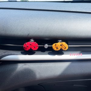 Könnte beinhalten: Zwei gehäkelte Auto-Lufterfrischer, einer rot und einer gelb, jeweils mit einer Ansammlung von drei Kugeln. Die Erfrischer sind an einer silbernen Lüftung befestigt. Der Text "XIN-H-VN" ist sichtbar.