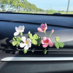 May include: Crocheted car vent clip featuring white and pink flowers with green leaves. The flowers are attached to a black clip, designed to fit into a car's air vent. The text "XINHVN" is visible in pink.
