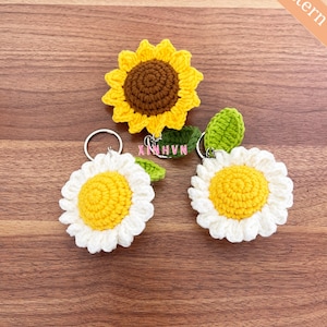 Op de afbeelding: Drie gebreide bloemen sleutelhangers. Een zonnebloem sleutelhanger met een bruin centrum en gele bloemblaadjes. Twee madeliefjes sleutelhangers met witte bloemblaadjes en gele centra. De sleutelhangers liggen op een houten oppervlak. De tekst "PDF Pattern" staat in de rechterbovenhoek van de afbeelding.