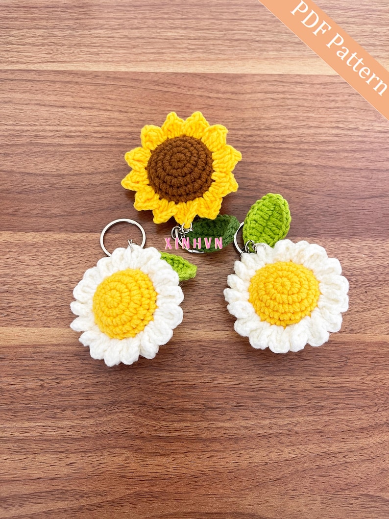Op de afbeelding: Drie gebreide bloemen sleutelhangers. Een zonnebloem sleutelhanger met een bruin centrum en gele bloemblaadjes. Twee madeliefjes sleutelhangers met witte bloemblaadjes en gele centra. De sleutelhangers liggen op een houten oppervlak. De tekst "PDF Pattern" staat in de rechterbovenhoek van de afbeelding.