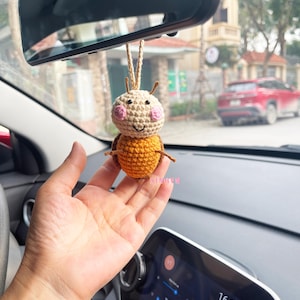 May include: A crocheted bee car charm with a yellow and orange body, brown eyes, and pink cheeks. The bee is hanging from a rearview mirror in a car.