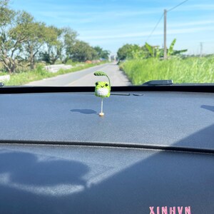 May include: A small, crocheted green frog with a leaf on its head, mounted on a thin, gold-colored stick. The frog is positioned on a dark dashboard, with the text "XINHVN" in pink letters. The background shows a road and a bright blue sky.