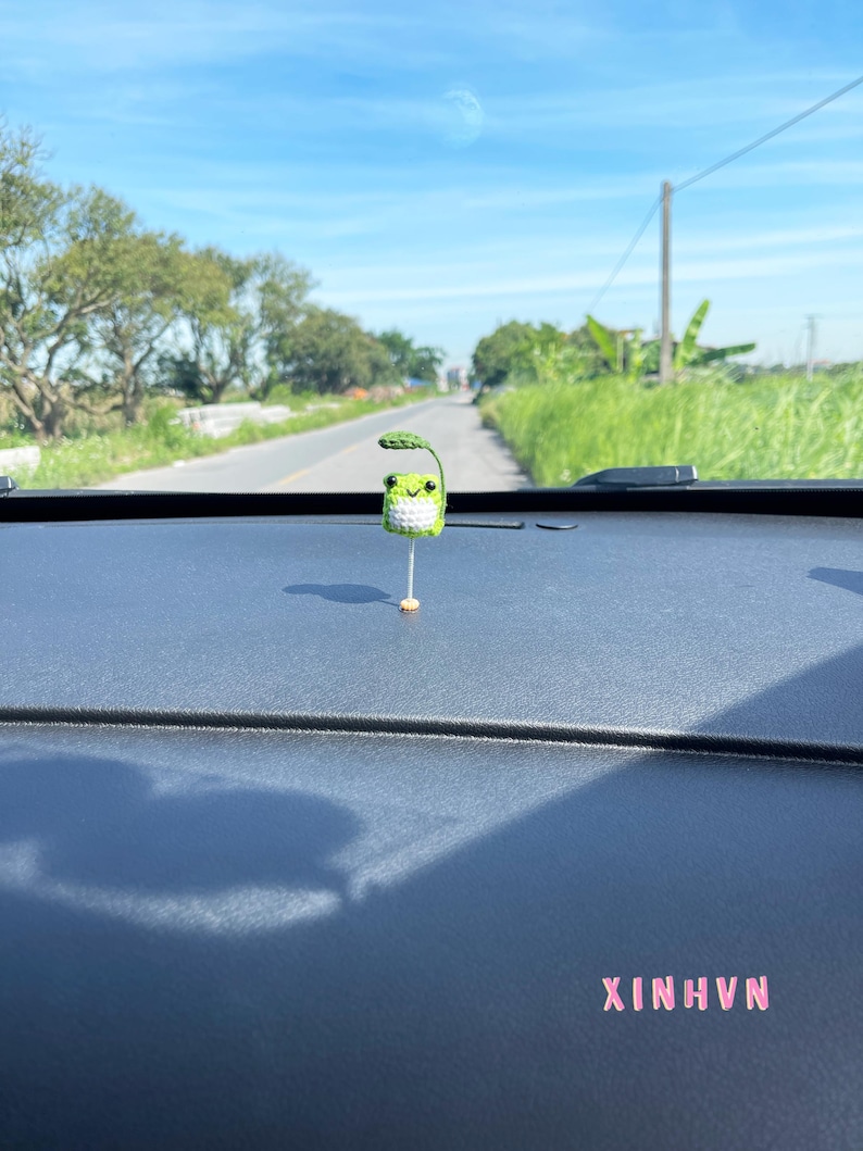 May include: A small, crocheted green frog with a leaf on its head, mounted on a thin, gold-colored stick. The frog is positioned on a dark dashboard, with the text "XINHVN" in pink letters. The background shows a road and a bright blue sky.