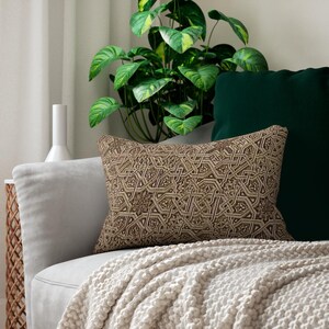 May include: A decorative rectangular pillow with a detailed geometric pattern in shades of brown and beige. The pillow is placed on a light gray sofa, with a green pillow and a white blanket. A houseplant is in the background.