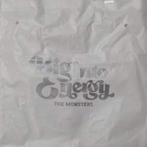 Op de afbeelding: Een witte plastic boodschappentas met een gekreukelde textuur en twee uitgestanste handvatten aan de bovenkant. De tas is voorzien van zilverkleurige metallic tekst met de woorden Big into Energy en THE MONSTERS in een vet, gestileerd lettertype.