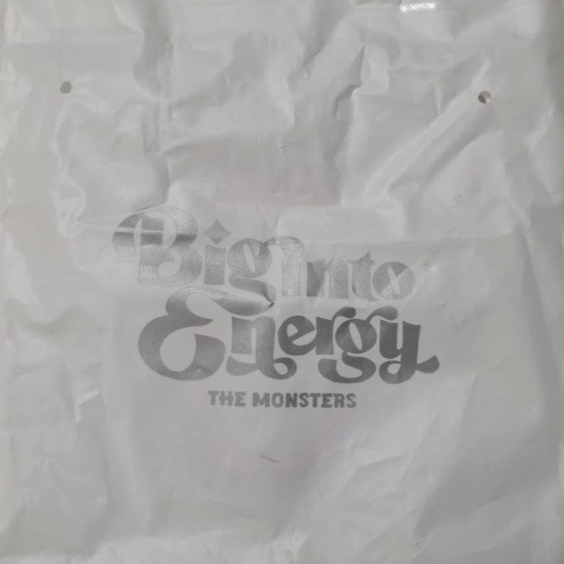 Op de afbeelding: Een witte plastic boodschappentas met een gekreukelde textuur en twee uitgestanste handvatten aan de bovenkant. De tas is voorzien van zilverkleurige metallic tekst met de woorden Big into Energy en THE MONSTERS in een vet, gestileerd lettertype.