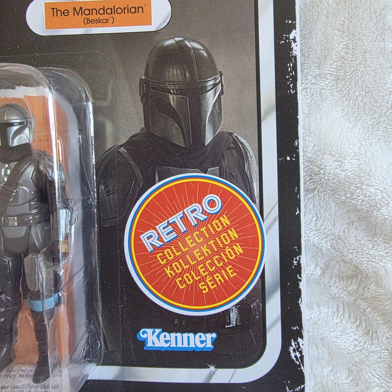 May include: A retro-style action figure of The Mandalorian (Beskar) in its original packaging. The figure is in a dark gray suit with a helmet. The packaging includes a "Retro Collection" sticker and the Kenner logo.