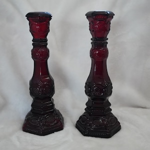 May include: A pair of deep red glass candle holders featuring a pressed pattern with circular and geometric motifs. Each candle holder has a wide base, a slender stem, and a flared top rim, showcasing a consistent dark ruby colour throughout.