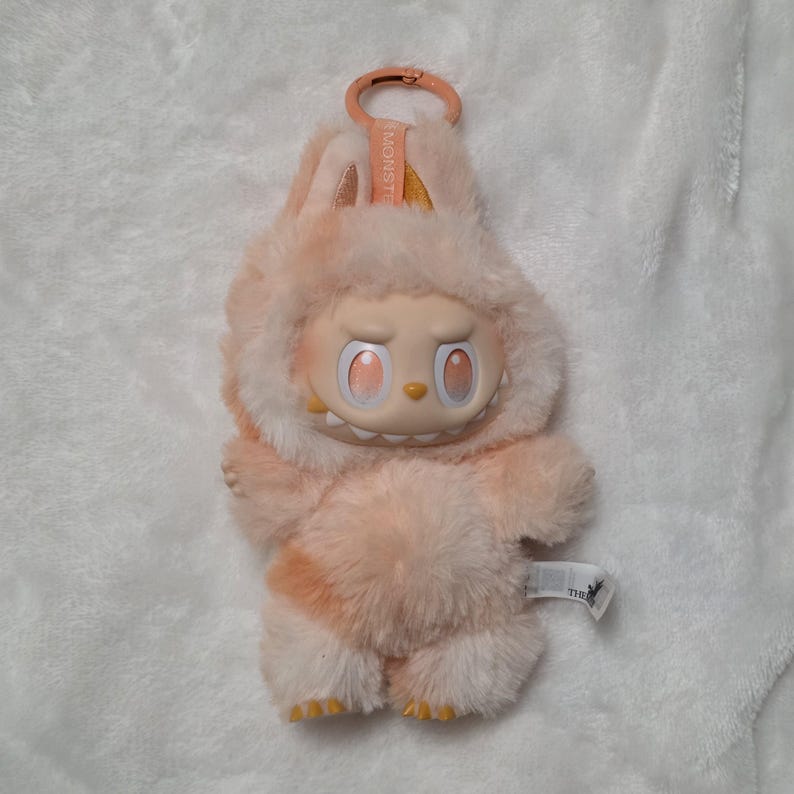 Op de afbeelding: Een knuffel met een perzikkleurig, pluizig lichaam en een plastic gezicht met grote, oranje getinte ogen, een kleine neus en zichtbare tanden. Het wezen heeft een capuchon met kleine oortjes en een plastic clip met het opschrift MONSTER.