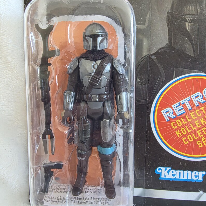 May include: A Mandalorian action figure in a clear plastic package. The figure is clad in silver and black armor, with a helmet and a dark scarf. Accessories include a staff and a blaster. The packaging features the word "RETRO" and the Kenner logo.