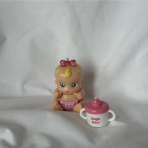 May include: A small plastic baby doll with blonde hair, a red bow, and a pink polka-dot diaper, accompanied by a white sippy cup with a pink lid and handles, featuring the text Grape Juice printed in pink on the front of the cup.