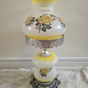 May include: A hurricane lamp featuring a white and yellow ombre glass shade and base, both decorated with yellow rose floral patterns. The lamp includes a silver-toned metal filigree gallery and a decorative silver-toned metal base with a power cord.