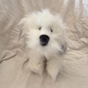 Puede incluir: Un juguete de peluche blanco con forma de perro, de aspecto esponjoso y desgreñado. El juguete tiene una nariz negra y ojos negros pequeños. Las orejas tienen un toque de gris. El perro está sentado, con las patas delanteras visibles.